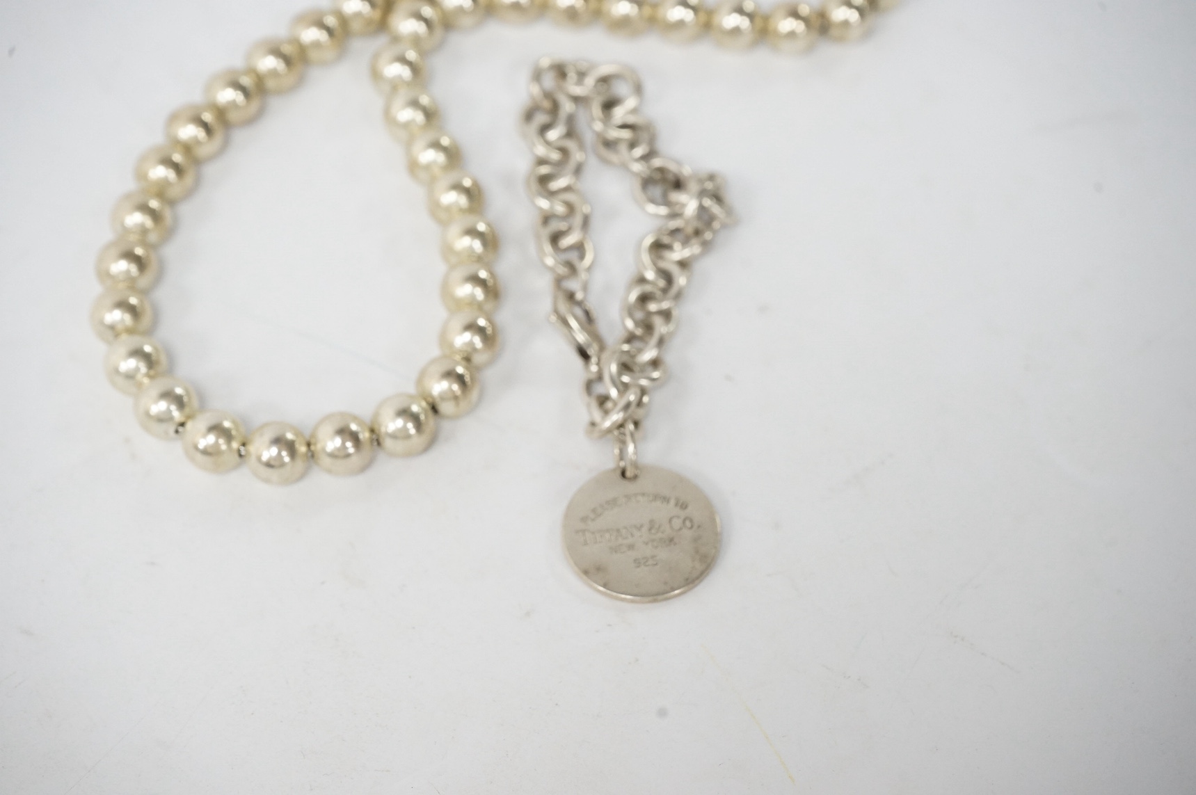 Tiffany & Co., a bracelet and necklace, the necklace composed of silver beads on a silver chain, length 45cm; the bracelet of circular linking suspending a medallion pendant inscribed 'Please return to Tiffany & Co.', le